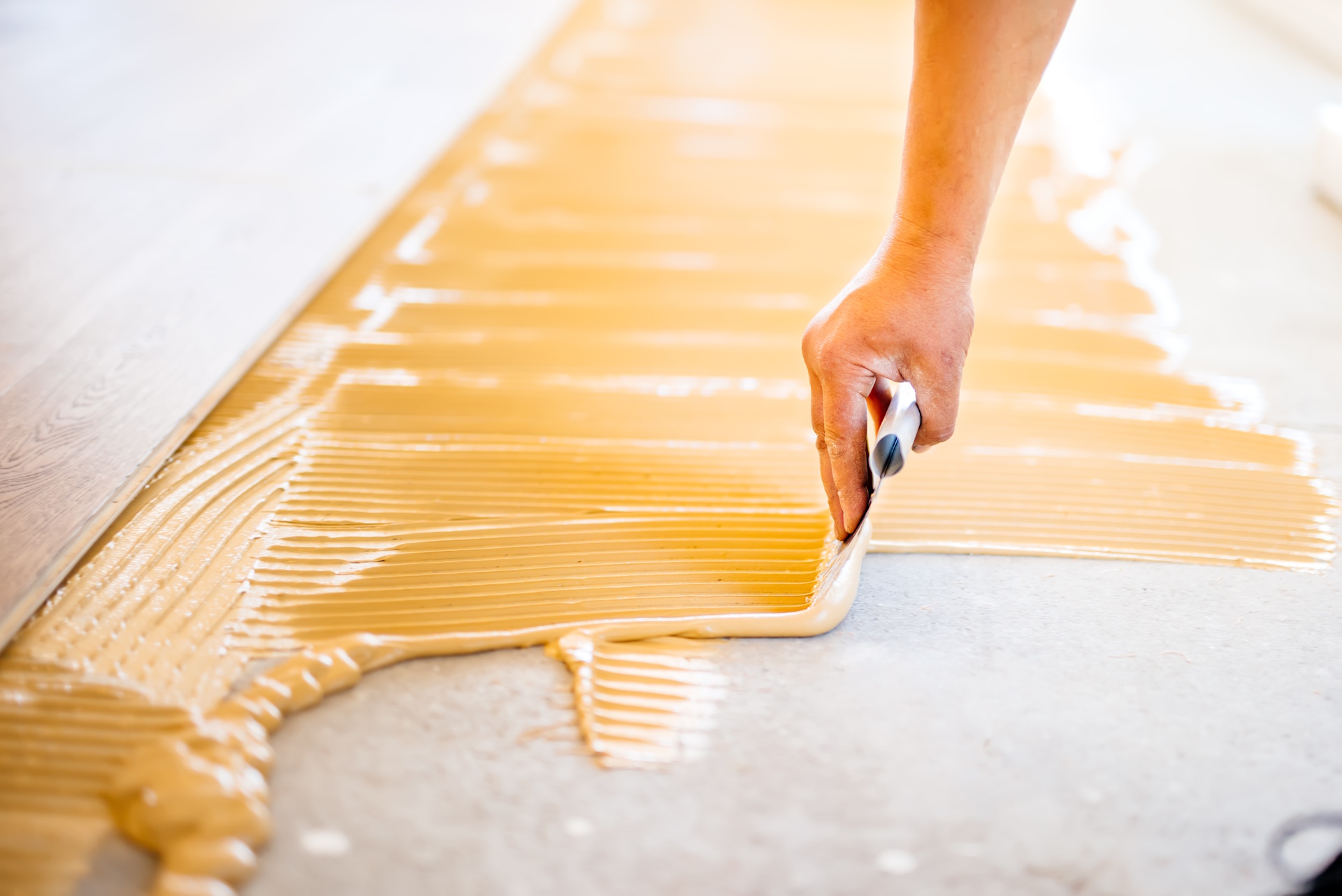 Ein Parkettboden sollte immer fachgerecht verlegt werden, um Langlebigkeit, Stabilität und ein perfektes Erscheinungsbild zu garantieren. Professionelle Handwerker sorgen für die richtige Vorbereitung des Untergrunds, die präzise Verklebung und ein makelloses Finish, das Wohnräume aufwertet.  Bildbeschreibung: Das Bild zeigt eine Nahaufnahme einer Hand, die mit einer Zahnspachtel Kleber auf einem Boden verteilt. Im oberen Bildbereich ist bereits ein Teil eines Holzbodens verlegt. Die Szene zeigt den handwerklichen Vorgang beim Verkleben eines Parkettbodens auf einem Untergrund.